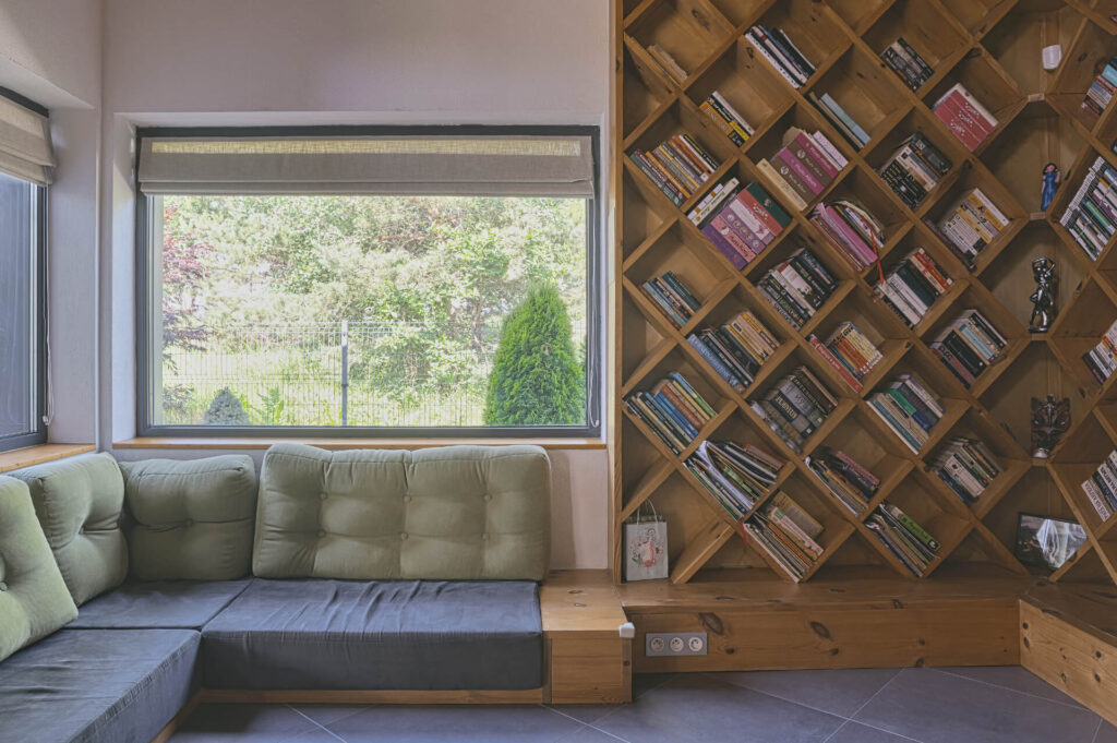 Модерна дневна от интериорен дизайн Studio28a със зелена секция и диагонална дървена етажерка за книги до голям прозорец.