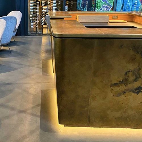 Модерен бар плот с бронзово покритие от Интериорен дизайн Studio28a, подсветка и рафтове с бутилки вино зад него.