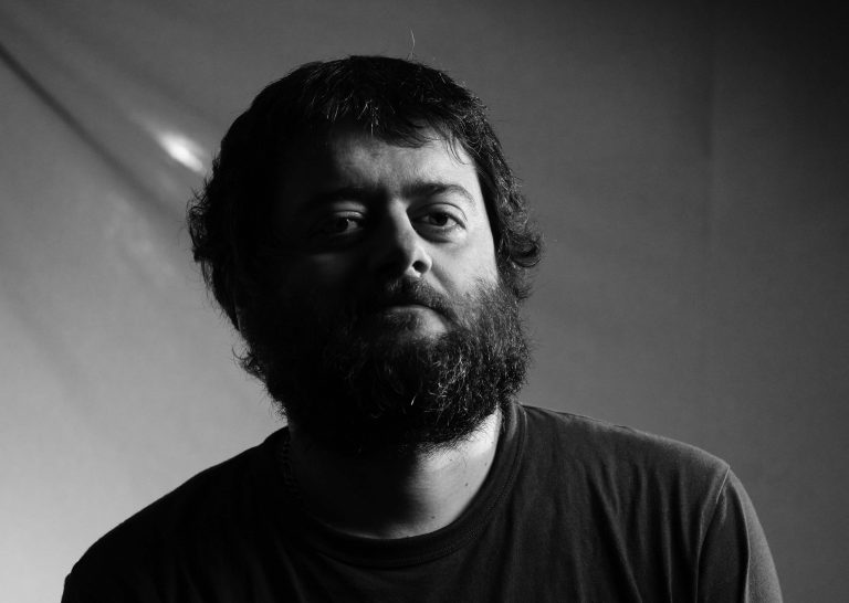 Мъж с брада и къдрава коса седи в частична сянка, оформен от Интериорен дизайн Studio28a, в черно-бял портрет.