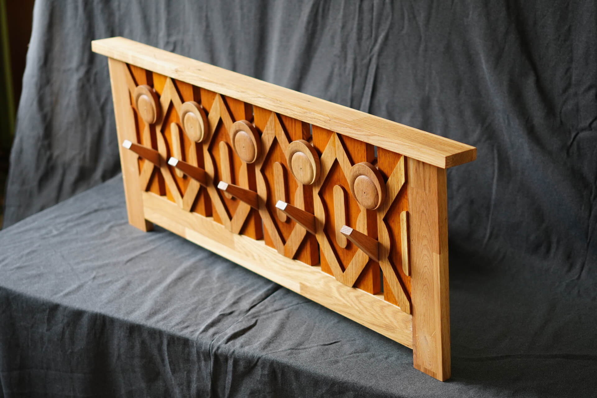 Дървена закачалка за дрехи с пет копчета и зигзагообразна шарка, изработена от Интериорен дизайн Studio28a, на фона на тъмен плат.