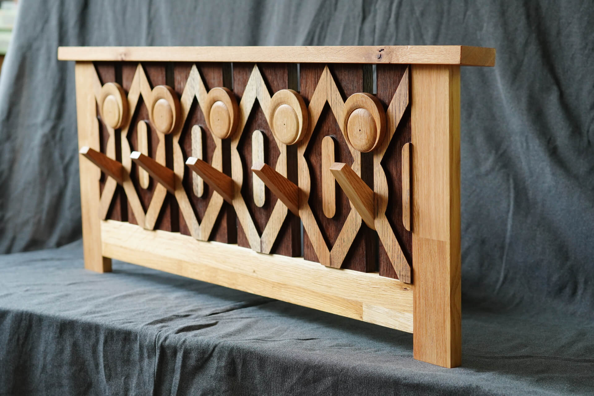 Дървена закачалка за дрехи с геометрични шарки от Интериорен дизайн Studio28a, поставена на фона на драпиран сив плат.