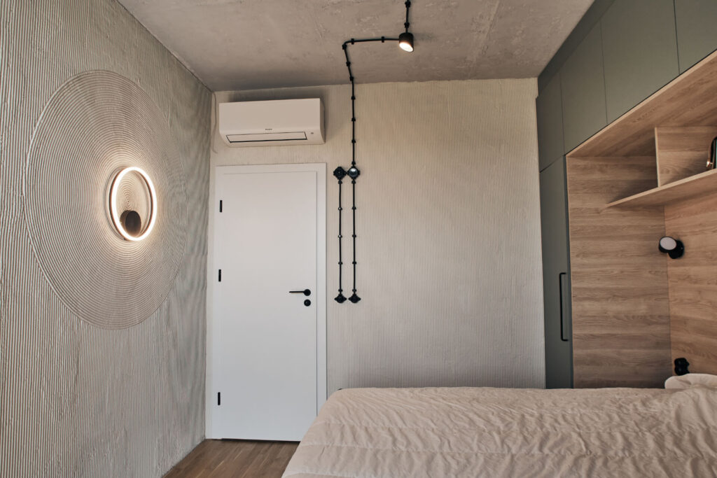 Минималистична спалня от Интериорен дизайн Studio28a с текстурирани стени, модерно осветление, дървени акценти и климатик над бяла врата.
