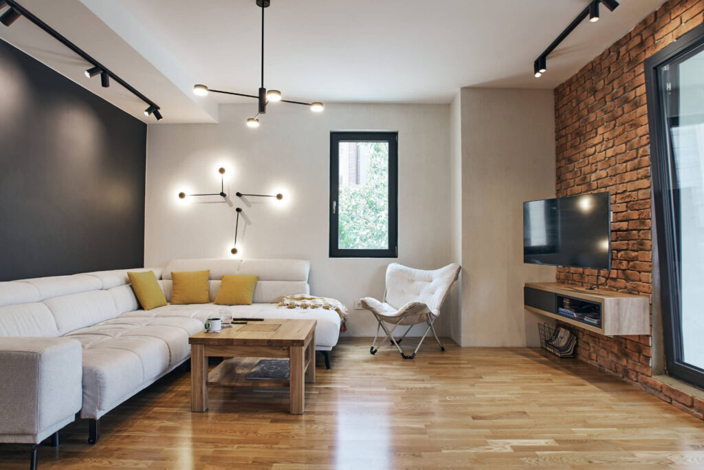 Модерна дневна от Интериорен дизайн Studio28a с бяла секция, дървена маса, телевизор и открита тухлена стена.