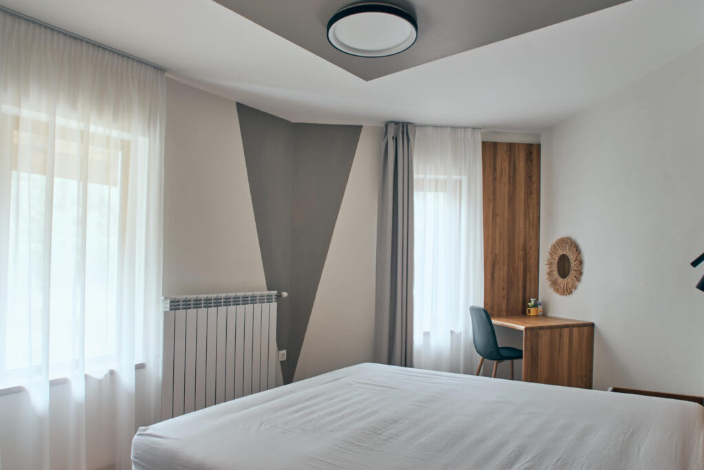 Минималистична спалня от Интериорен дизайн Studio28a с прозрачни завеси, дървено бюро и геометричен дизайн на стените.