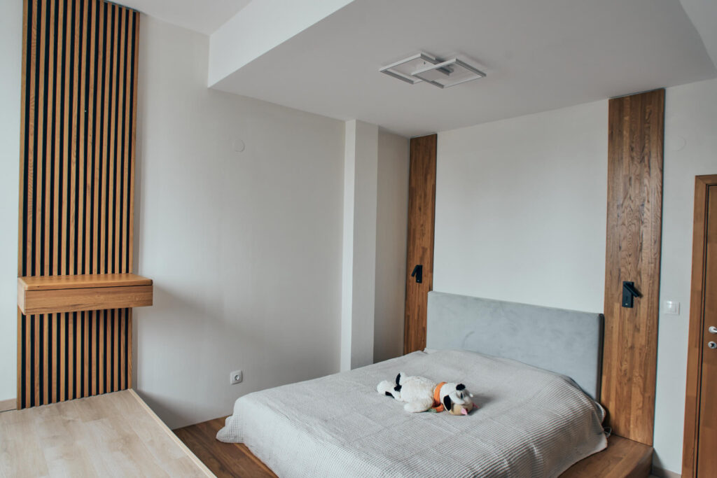 Модерна спалня с дървена акцентна стена, семпло легло и плюшена кучешка играчка от Интериорен дизайн Studio28a.