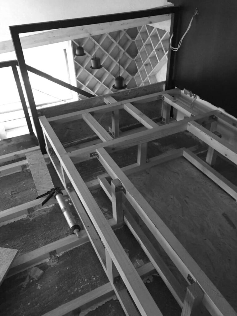 Незавършена дървена рамка на платформа в процес на изграждане от Интериорен дизайн Studio28a, висящи лампи и решетъчна стена отзад.